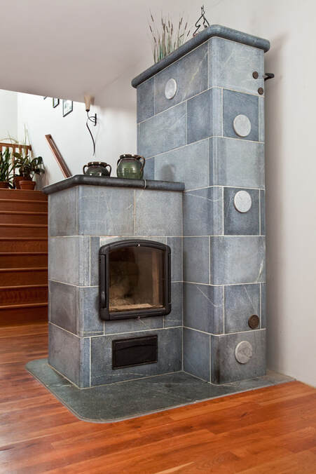 A grey NEHS masonry heater near a stairwell. The heater has a glass door and two decorative pots on top of it. 